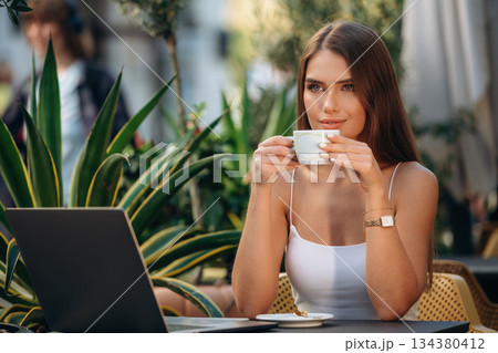Fresh hot drink in cup. Beautiful young woman is outdoors of the restaurant by the table 134380412