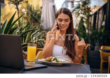 Talking by phone. Beautiful young woman is outdoors of the restaurant by the table Talking by phone. Beautiful young woman is outdoors of the restaurant by the table 134380504