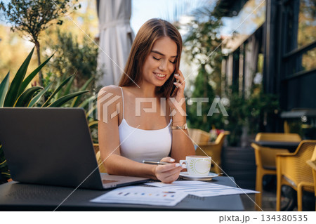 Phone call, with documents. Beautiful young woman is outdoors of the restaurant by the table Phone call, with documents. Beautiful young woman is outdoors of the restaurant by the table 134380535