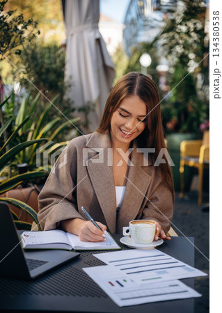Remote work, with drink and documents. Beautiful young woman is outdoors of the restaurant by the table Remote work, with drink and documents. Beautiful young woman is outdoors of the restaurant by the table 134380538