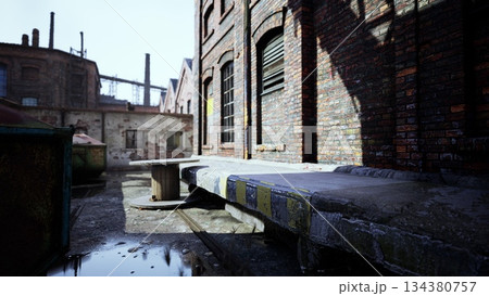 A narrow urban alleyway features old brick buildings with black windows and a wooden spool table. Sunlight casts shadows on the wet ground, highlighting the industrial charm of the space. 134380757