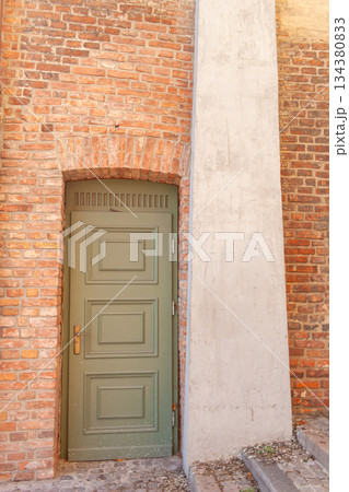 Green door set in a brick wall with concrete structure in the background 134380833