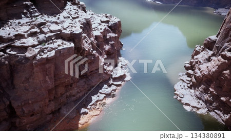 Beneath the towering canyon cliffs, a tranquil river glimmers under the warm sun. Smooth stones line the sandy banks, creating a peaceful natural scene. The blue water reflects the clear sky above. Beneath the towering canyon cliffs, a tranquil river glimmers under the warm sun. Smooth stones line the sandy banks, creating a peaceful natural scene. The blue water reflects the clear sky above. 134380891