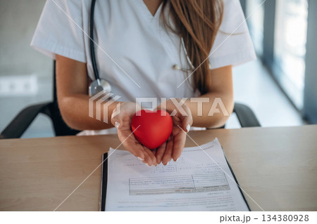 Close up view, holding little red heart. Professional female doctor in the clinic indoors 134380928