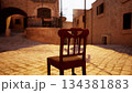A solitary wooden chair is placed in a quiet, sunlit cobblestone street. The warm glow of sunset creates a tranquil atmosphere, inviting reflection in a historic town setting. 134381883