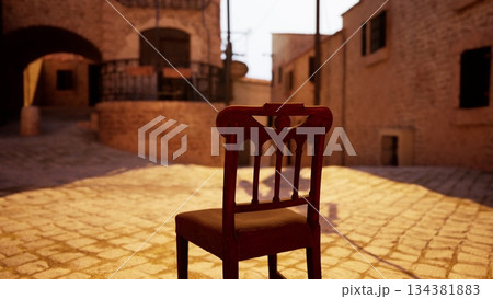A solitary wooden chair is placed in a quiet, sunlit cobblestone street. The warm glow of sunset creates a tranquil atmosphere, inviting reflection in a historic town setting. 134381883