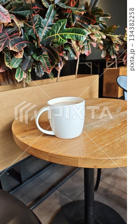 White ceramic coffee mug placed on a round wooden table inside a modern cafe with decorative plants in the background. Concept of calm coffee break, minimalism and cozy cafe atmosphere 134382258