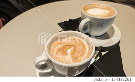 Two cups of cappuccino with creamy milk foam served on a round cafe table with spoons and napkins. Concept of shared coffee moment, social interaction and relaxed cafe lifestyle Two cups of cappuccino with creamy milk foam served on a round cafe table with spoons and napkins. Concept of shared coffee moment, social interaction and relaxed cafe lifestyle 134382261
