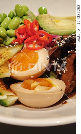 Close-up of Asian style bowl with soft boiled egg, edamame, avocado, chili pepper and glazed meat on a white plate. Concept of healthy fusion cuisine, balanced nutrition and modern comfort food Close-up of Asian style bowl with soft boiled egg, edamame, avocado, chili pepper and glazed meat on a white plate. Concept of healthy fusion cuisine, balanced nutrition and modern comfort food 134382262
