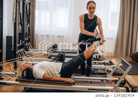 Laying down and doing legs exercise. Female trainer is helping old woman 134382454