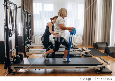 Blue colored resistance loops in hands for the legs. Female trainer is helping old woman to exercise Blue colored resistance loops in hands for the legs. Female trainer is helping old woman to exercise 134382505