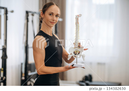 Showing the bones of the model of spine. Young woman is in the gym with training equipment 134382630