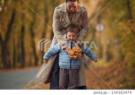 Fun times, behind the boy. Mother with her son are having a walk in autumn forest near the road Fun times, behind the boy. Mother with her son are having a walk in autumn forest near the road 134383474