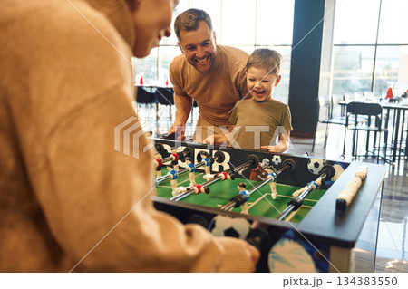 Smiling, enjoying. Family of dad, mother and son that are playing table football game indoors Smiling, enjoying. Family of dad, mother and son that are playing table football game indoors 134383550