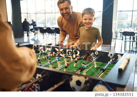 Smiling, enjoying. Family of dad, mother and son that are playing table football game indoors Smiling, enjoying. Family of dad, mother and son that are playing table football game indoors 134383551