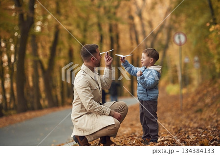 Father and son are playing with paper plane in the autumn forest near the road Father and son are playing with paper plane in the autumn forest near the road 134384133