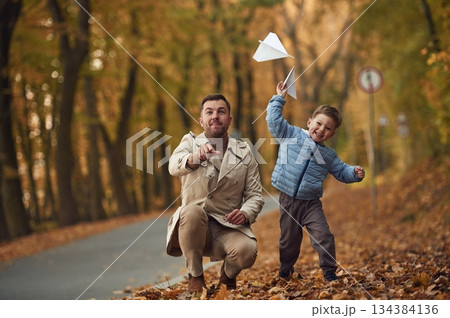 Father and son are playing with paper plane in the autumn forest near the road Father and son are playing with paper plane in the autumn forest near the road 134384136