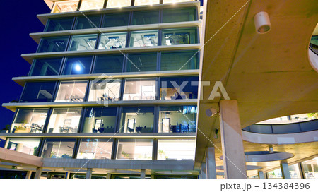 Office building in the evening with lightened workspaces. Modern, glazed skyscraper in financial district. Late hours in the office. 134384396