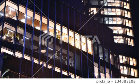 Modern office building in city center illuminated at night. Rows of lit windows against the architectural grid of a modern facade. Concrete and glass facade in its night version. 134385108