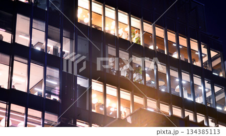 Modern office building in city center illuminated at night. Rows of lit windows against the architectural grid of a modern facade. Concrete and glass facade in its night version. 134385113
