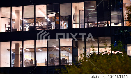 Modern office building in city center illuminated at night. Rows of lit windows against the architectural grid of a modern facade. Concrete and glass facade in its night version. 134385133