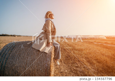 Woman in a Trench Coat Sitting on a Hay Bale in a Field Woman in a Trench Coat Sitting on a Hay Bale in a Field 134387796