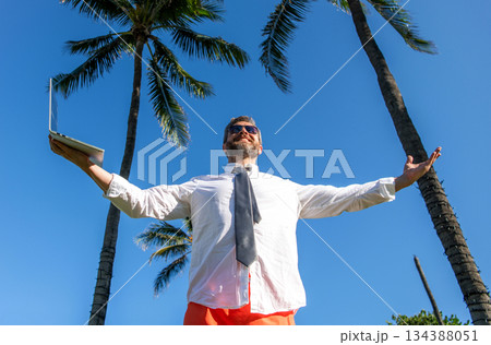 Businessman on summer vacation. Online freelance work. Businessman boss with laptop. Business man using laptop near palm tree. Business trip. Man freelance work online at beach. Happy holiday 134388051