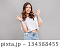 Beautiful teenage girl with wavy brown hair smiling and raising hands in playful gesture, posing in studio on gray background, young female fashion model in casual white top and denim 134388455
