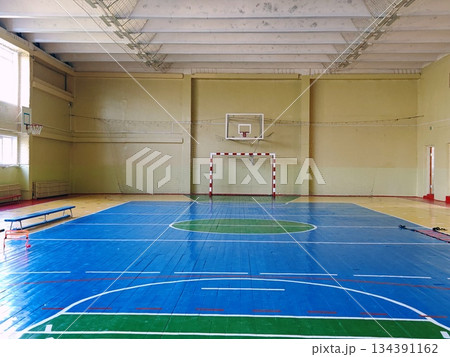 Empty School Gymnasium with Basketball and Handball Court Markings. Spacious empty indoor school sports hall with blue wooden floor, basketball hoops and handball goal. Concept of education, sport Empty School Gymnasium with Basketball and Handball Court Markings. Spacious empty indoor school sports hall with blue wooden floor, basketball hoops and handball goal. Concept of education, sport 134391162