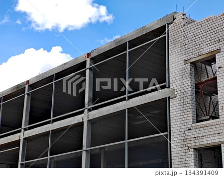Unfinished Multi-Story Building Under Construction. Modern multi-floor concrete building under construction with exposed floors and brick walls against blue sky. Unfinished Multi-Story Building Under Construction. Modern multi-floor concrete building under construction with exposed floors and brick walls against blue sky. 134391409