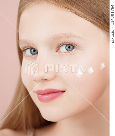 Portrait of a smiling cheerful girl applying face cream on cheek for skin glow. Advertising face cream and skincare products for teens. Blonde girl with blue eyes isolated in studio on pink background 134391764