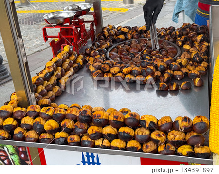 Roasted chestnuts arranged on a metal tray at a street food stand. Seasonal street cuisine, traditional roasting methods, and warm comfort food associated with autumn markets. Roasted chestnuts arranged on a metal tray at a street food stand. Seasonal street cuisine, traditional roasting methods, and warm comfort food associated with autumn markets. 134391897
