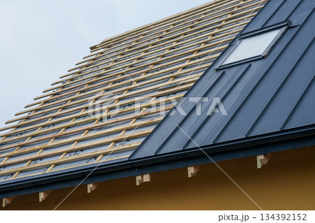 New house roof under construction with wooden battens and modern metal covering around attic window New house roof under construction with wooden battens and modern metal covering around attic window 134392152