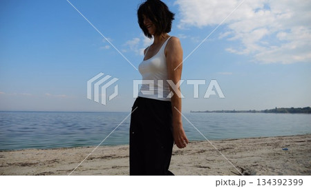 Happy adult woman going on sandy beach near shoreline at sunny day. Beautiful lady walking on seashore with calm waves at background. Concept of summer vacation or holiday. Slow motion 134392399
