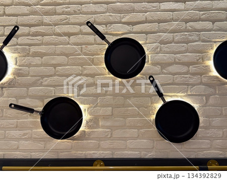 Decorative wall installation of black frying pans mounted on white brick background with accent lighting. Culinary theme, modern interior design, playful composition, and restaurant atmosphere through 134392829