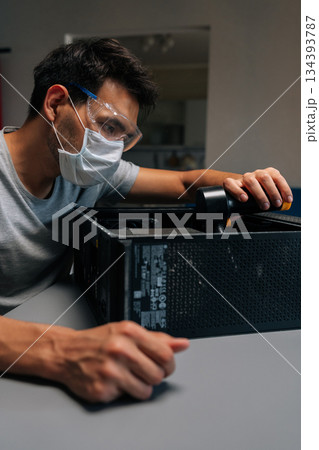 Vertical portrait of computer technician in protective mask and safety eyewear cleaning system unit with vacuum during precision hardware maintenance. Servicing motherboard of dusty computer. Vertical portrait of computer technician in protective mask and safety eyewear cleaning system unit with vacuum during precision hardware maintenance. Servicing motherboard of dusty computer. 134393787