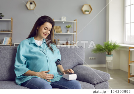 Happy healthy young pregnant woman sitting on sofa and measuring her blood pressure Happy healthy young pregnant woman sitting on sofa and measuring her blood pressure 134393993