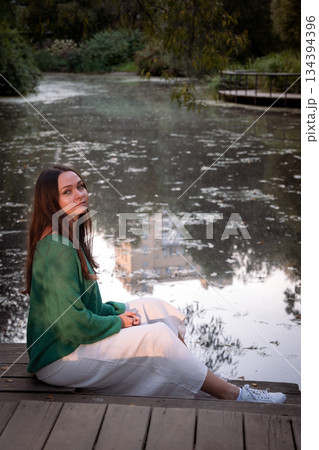 Young woman sits on a wooden deck by the water, looking down, soft natural light, muted green and gray tones, quiet and introspective mood 134394396