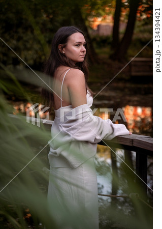 Young woman in a dress, cardigan stands on a wooden bridge by a pond, looking to the side, soft daylight, muted green tones, calm, introspective mood 134394421