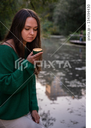 A woman holds a small lit candle in her hands, soft flame reflecting on her face against a blurred water and greenery background A woman holds a small lit candle in her hands, soft flame reflecting on her face against a blurred water and greenery background 134394430