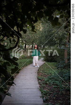 Woman in a green sweater and light skirt walks along a wooden boardwalk surrounded by dense trees and greenery, creating a calm and natural atmosphere Woman in a green sweater and light skirt walks along a wooden boardwalk surrounded by dense trees and greenery, creating a calm and natural atmosphere 134396466