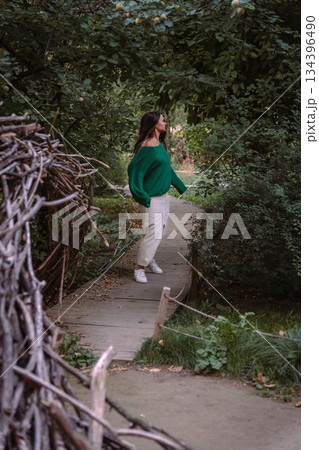 A woman in a green sweater and light skirt stands on a narrow wooden path surrounded by dense greenery, creating a quiet and reflective outdoor scene A woman in a green sweater and light skirt stands on a narrow wooden path surrounded by dense greenery, creating a quiet and reflective outdoor scene 134396490
