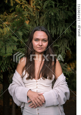 Young woman in a light dress and cardigan stands against evergreen branches, soft daylight, deep green tones, calm and confident mood Young woman in a light dress and cardigan stands against evergreen branches, soft daylight, deep green tones, calm and confident mood 134396551