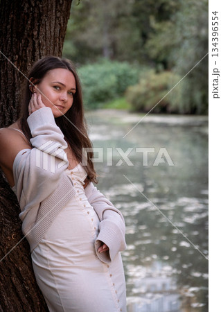 Young woman in a light dress and cardigan stands by a river near a tree, touching her face, soft daylight, muted green tones, calm and intimate mood 134396554