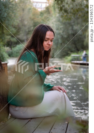 Young woman in a green sweater holds a small lit candle while sitting by a calm pond, soft daylight, muted green tones, quiet and contemplative mood Young woman in a green sweater holds a small lit candle while sitting by a calm pond, soft daylight, muted green tones, quiet and contemplative mood 134396572