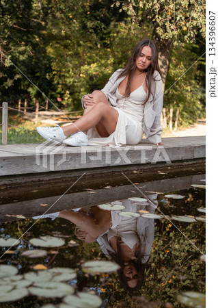 Young woman in a light dress sits on a wooden edge by a calm pond with reflections and floating leaves, soft daylight, warm natural tones, serene mood 134396607