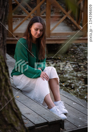 Young woman sits on a wooden boardwalk by a pond, fallen leaves on water, soft daylight, natural colors, calm and intimate mood Young woman sits on a wooden boardwalk by a pond, fallen leaves on water, soft daylight, natural colors, calm and intimate mood 134396609