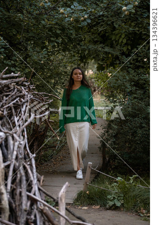 A woman in a green sweater and light skirt walks along a narrow wooden path surrounded by lush greenery, creating a calm and intimate nature scene 134396621