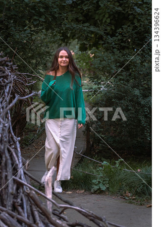 A woman in a green sweater and light skirt walks toward the camera along a narrow garden path, surrounded by dense foliage and natural textures A woman in a green sweater and light skirt walks toward the camera along a narrow garden path, surrounded by dense foliage and natural textures 134396624