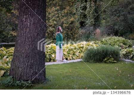A woman in a green sweater and light skirt stands in a lush garden, surrounded by tall ivy-covered trees, leafy plants, and soft natural light 134396676